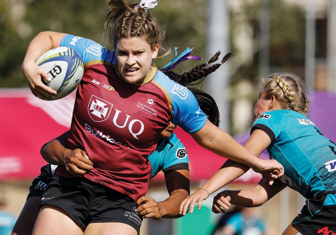 Student in blue and maroon UQ jersey holding the football and dodging a tackle in a game of women's rugby.