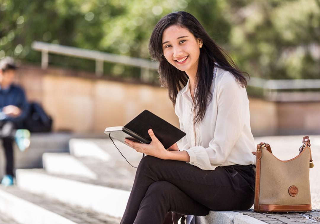 A young woman holding a tablet sits outside, smiling while looking at the camera
