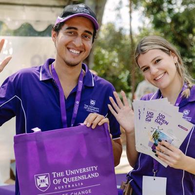 UQ Students in purple polo shirts hold leaflets and a UQ tote bag and wave at the camera