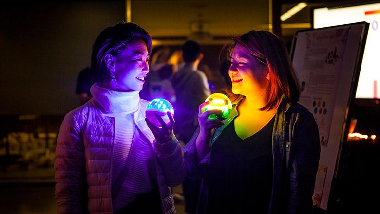 Two students holding multicoloured light balls. The rainbow of lights are reflecting off their faces.
