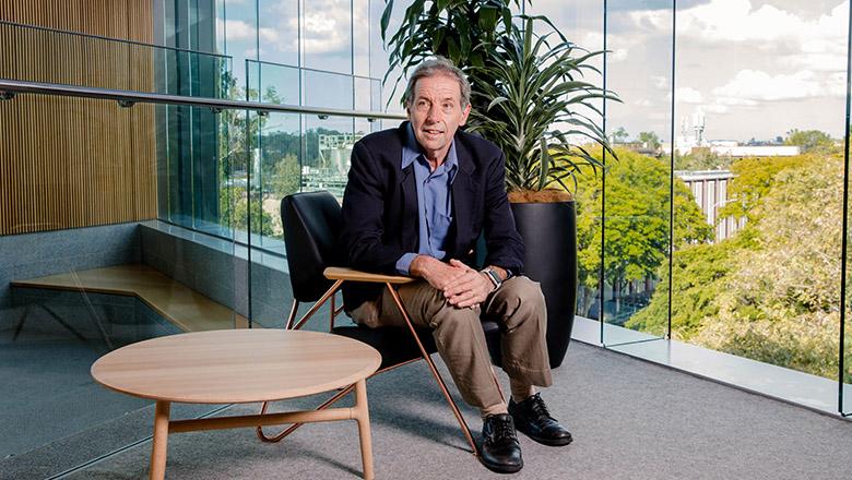 Professor John Quiggin sits on an armchair in front of a floor to ceiling window overlooking green trees
