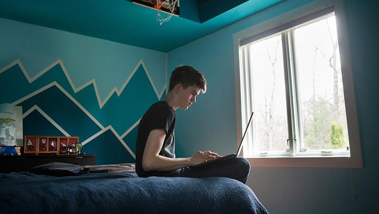 A teenager boy sits on his bed in his bedroom using his laptop