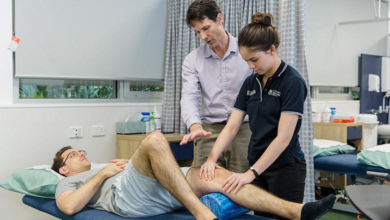 A UQ student examines the knee of a man lying on an examination table, with a supervisor guiding her