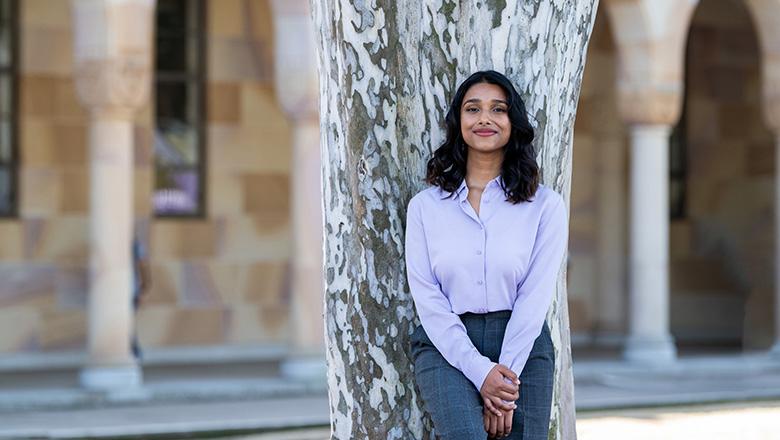 Nisha Gupta leans against a tree in UQ's Great Court with sandstone cloisters in the background