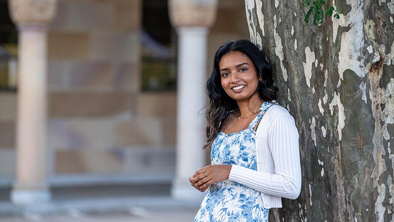 Chrisny Fernando, UQ Master of Public Health student leans against a tree in UQ's Great Court with sandstone buildings in the background