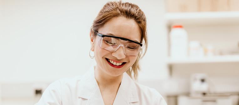 A woman wearing a lab coat and safety glasses working in a lab.