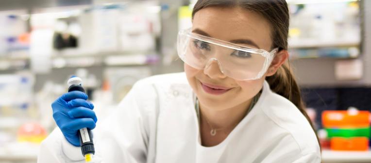 A student wearing safety glasses and a lab coat, using equipment in a lab.